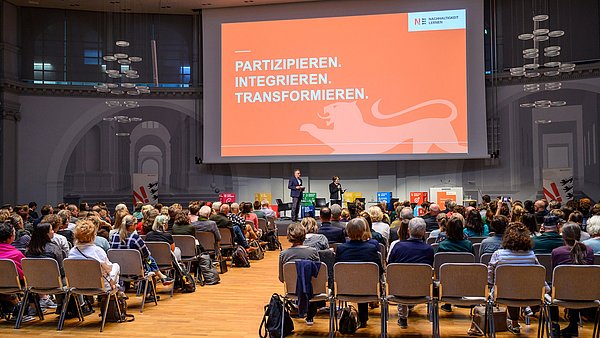 Blick in den vollbesetzten Veranstaltungssaal des BNE Events. Auf einem großen Präsentationsmonitor ist zu lesen: Partizipieren, integrieren, transformieren.