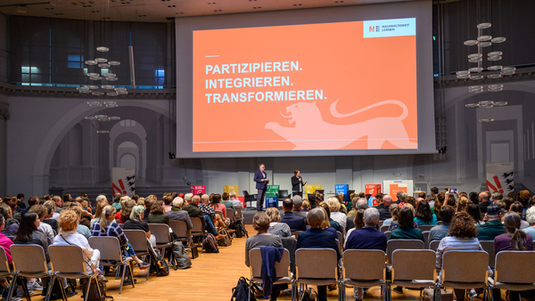 Blick in den vollbesetzten Veranstaltungssaal des BNE Events. Auf einem großen Präsentationsmonitor ist zu lesen: Partizipieren, integrieren, transformieren.