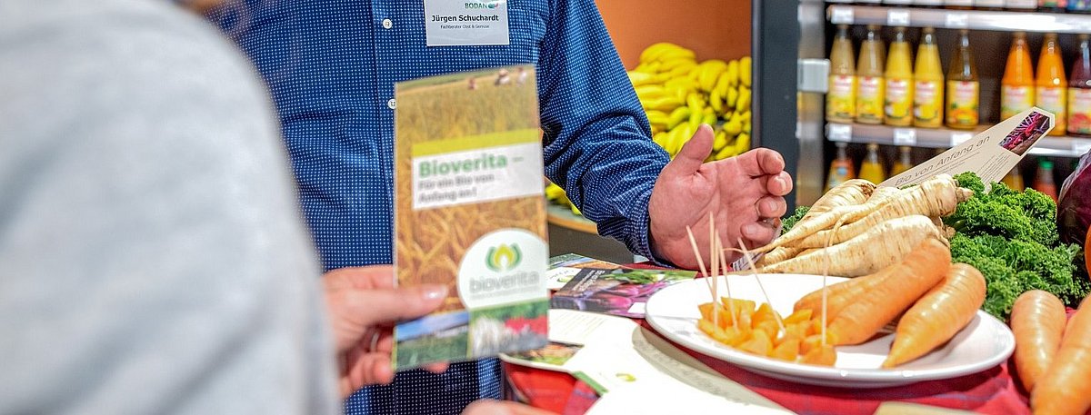 In einem Hofladen unterhalten sich zwei Personen, auf einem Tisch liegt frisches Gemüse und Käse