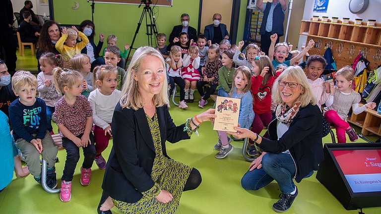 Thekla Walker MdL und eine Erzieherin zeigen den Preis in die Kamera. Im Hintergrund freuen sich die Kinder über den Gewinn