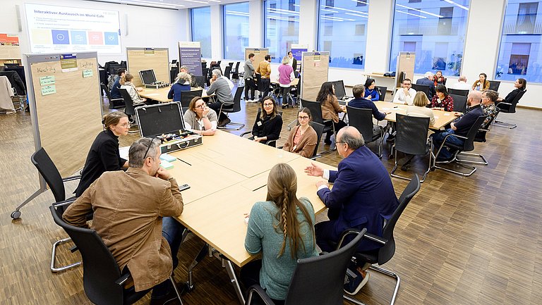 Um mehrere Tische sitzen viele Personen und tauschen sich in Diskussionen miteinerander aus.