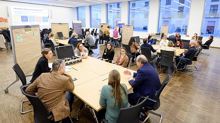 Um mehrere Tische sitzen viele Personen und tauschen sich in Diskussionen miteinerander aus.