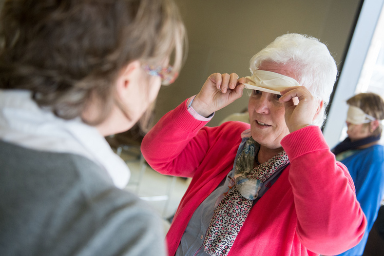 Eine ältere Dame in pinker Strickjacke lupft eine Augenbinde und blickt eine ihr gegenüberstehende Person an.