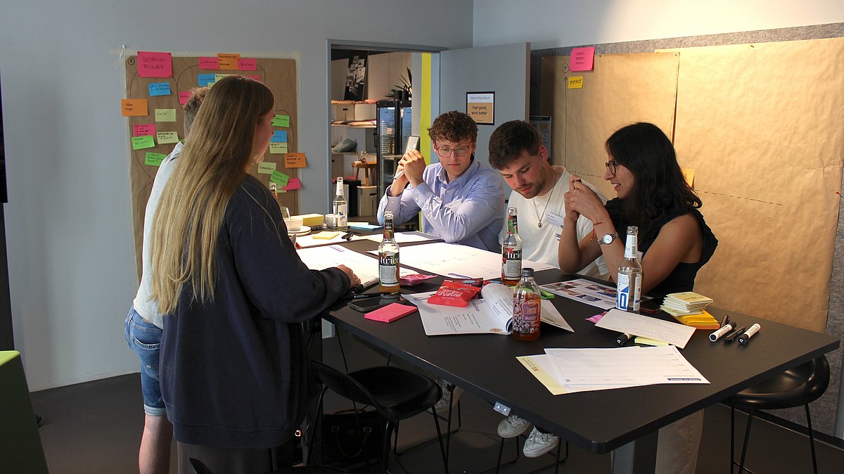 Fünf Personen stehen zentral im Bild um einen Tisch. Es liegen viele Zettel und Post-Its auf dem Tisch und es wird gemeinsam etwas bearbeitet.