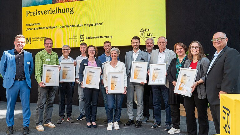 Farbige Fotografie: Gruppenbild auf der Bühne mit allen Preisträgerinnen und Preisträgern. Alle halten ihre Urkunden 'Sport & Nachhaltigkeit' in der Hand.