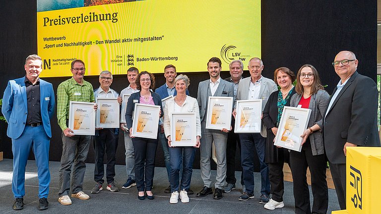 Farbige Fotografie: Gruppenbild auf der Bühne mit allen Preisträgerinnen und Preisträgern. Alle halten ihre Urkunden 'Sport & Nachhaltigkeit' in der Hand.