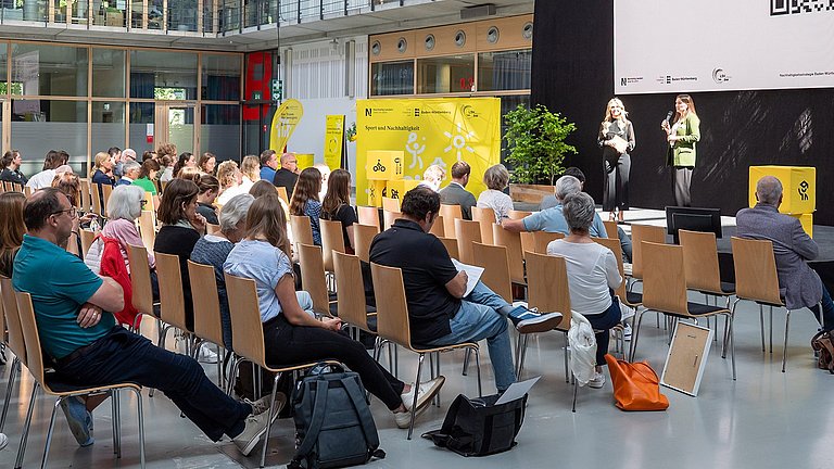 Farbige Fotografie: Zwei Frauen stehen auf einer Bühne vor großem QR-Code und sprechen beim Workshop-Hinweis 'Anmeldung zum Interessierten-Workshop am 22. Mai 2025'.