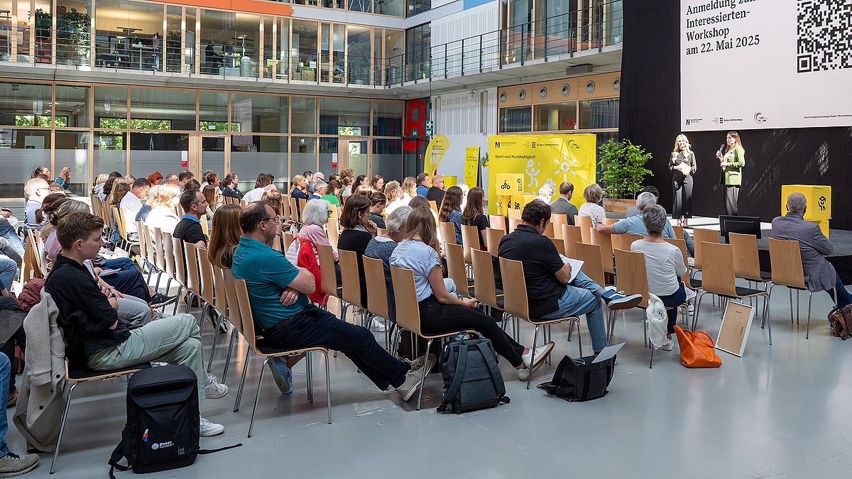 Farbige Fotografie: Zwei Frauen stehen auf einer Bühne vor großem QR-Code und sprechen beim Workshop-Hinweis 'Anmeldung zum Interessierten-Workshop am 22. Mai 2025'.