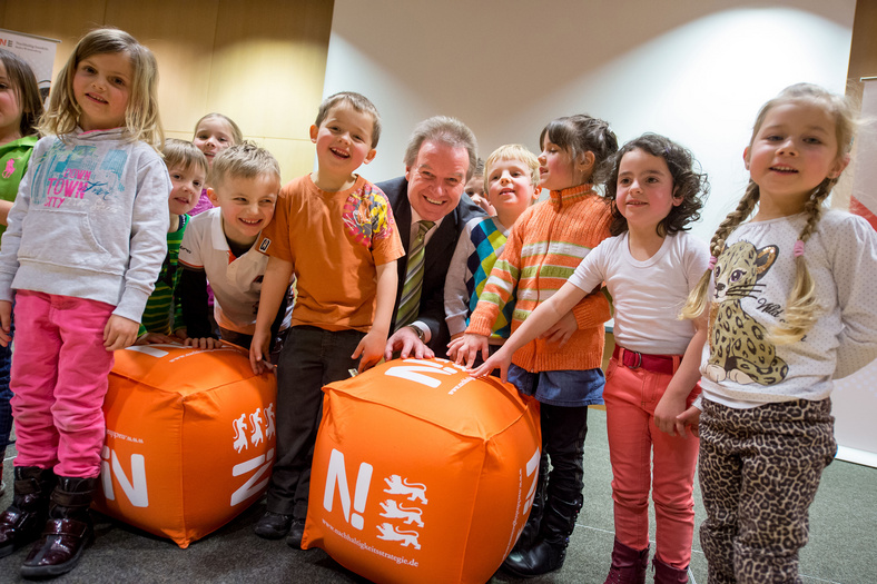 Gruppenfoto mit Minister Untersteller. Zehn Kinder im Kindergartenalter, stehend. Der Umweltminister in Hocke lacht in die Kamera. Im Vordergrund sind zwei orangefarbene Würfel mit dem Nachhaltigkeitslogo.