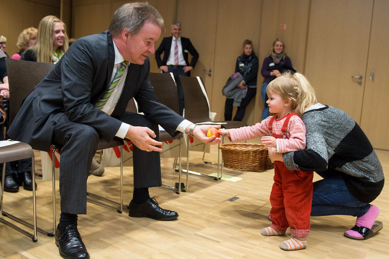 Ein ca. einjähriges Kind in roten Latzhosen, gestützt von einer Frau reicht dem sitzenden Minister Untersteller einen Apfel.