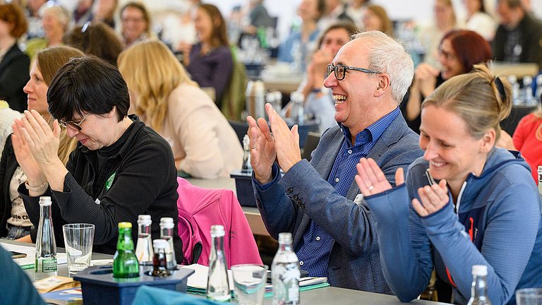 Mehrere Personen sitzen an Tischen mit Blick nach vorne gerichtet, klatschen in die Hände und lachen dabei.