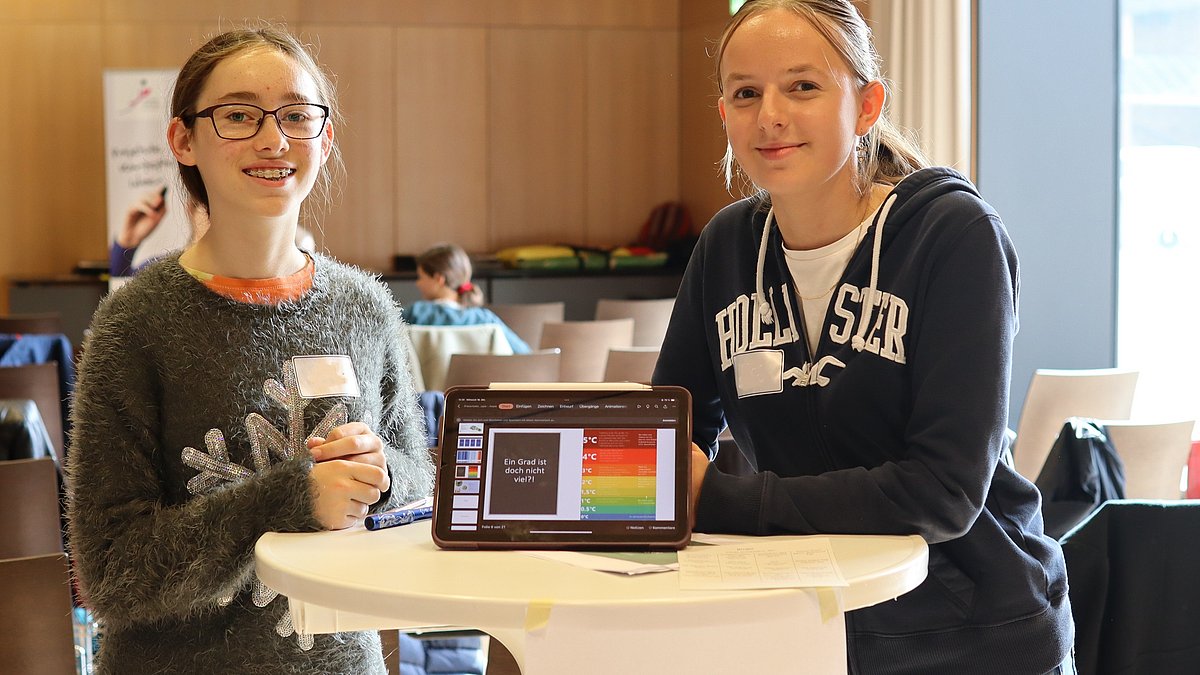 Zwei Jugendliche stehen um ein Tablet herum und lächeln, sie sollen Umweltmentorinnen darstellen.