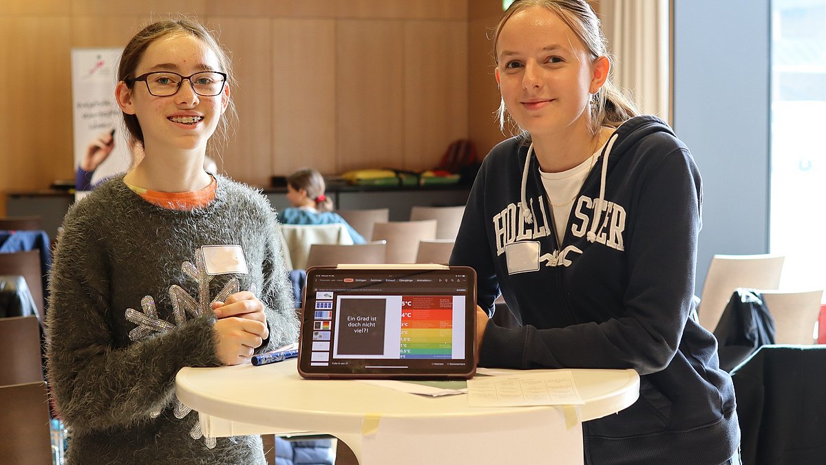 Zwei Jugendliche stehen um ein Tablet herum und lächeln, sie sollen Umweltmentorinnen darstellen.