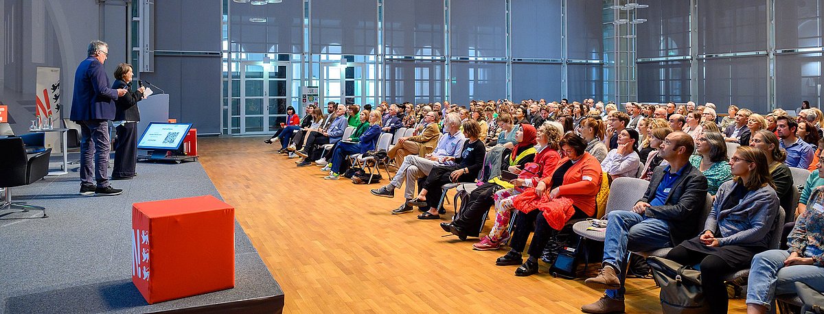 Links im Bild ist die Bühne zu sehen auf der die Moderatorin und der Moderator stehen und rechts die vollen Stuhlreihen während der Vormittagveranstaltung des BNE-BW.