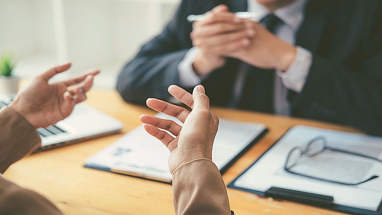 Zwei gestikulierende Hände über einem Tisch, auf der anderen Tischseite sitzt eine Person mit gefalteten Händen, deren Kopf nicht zu sehen ist. Auf dem Tisch liegen Unterlagen und ein Laptop.