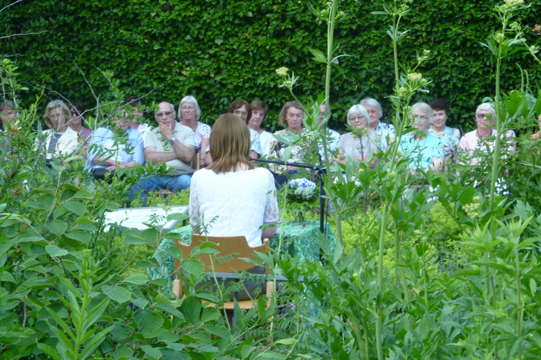 Autorin Susanne Rocholl liest vor einem Publikum inmitten eines grünen Blumenbeets aus ihrem Buch vor