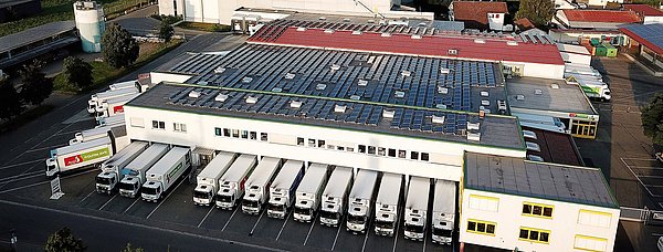 Eine Luftaufnahme des Firmengeländes der Früchte Jork GmbH. Auf den Dächern ist die Photovoltaik-Anlage erkennbar und im Hintergrund die Allgäuer Landschaft.