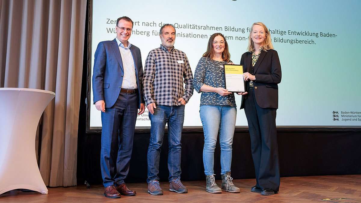 Umweltministerin Thekla Walker MdL und Kultusstaatssekretär Volker Schebesta MdL stehen zusammen mit einer Frau und einem Mann auf einer Bühne. Sie lächeln alle in die Kamera, die Umweltministerin und die Frau halten ein eingerahmtes BNE-Zertifikat.