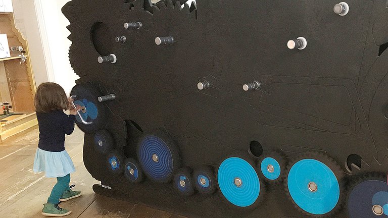 Ein Mädchen steht an einer Wand und dreht eine selbst zusammengestellte Zahnradkette.