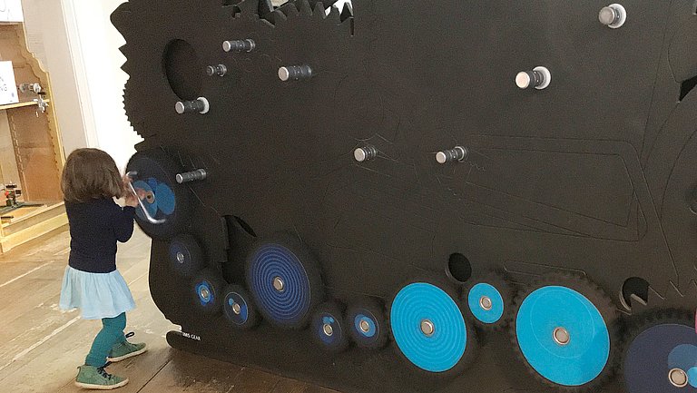 Ein Mädchen steht an einer Wand und dreht eine selbst zusammengestellte Zahnradkette.