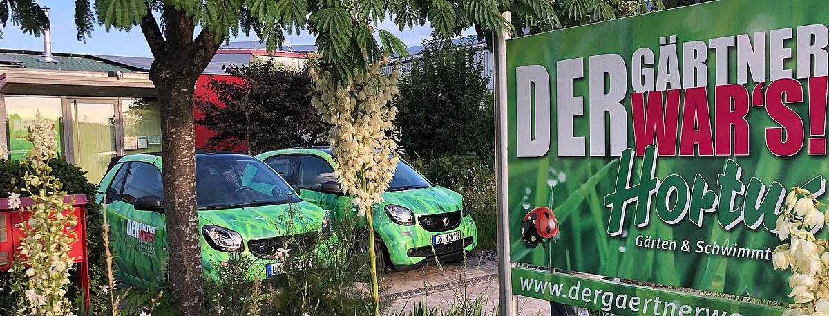 Ein Schild von Hortus mit der Beschriftung "Der Gärtner wars. Hortus". Im Hintergrund sind eine Gartenanlage und zwei grüne Smart-PKW zu sehen.