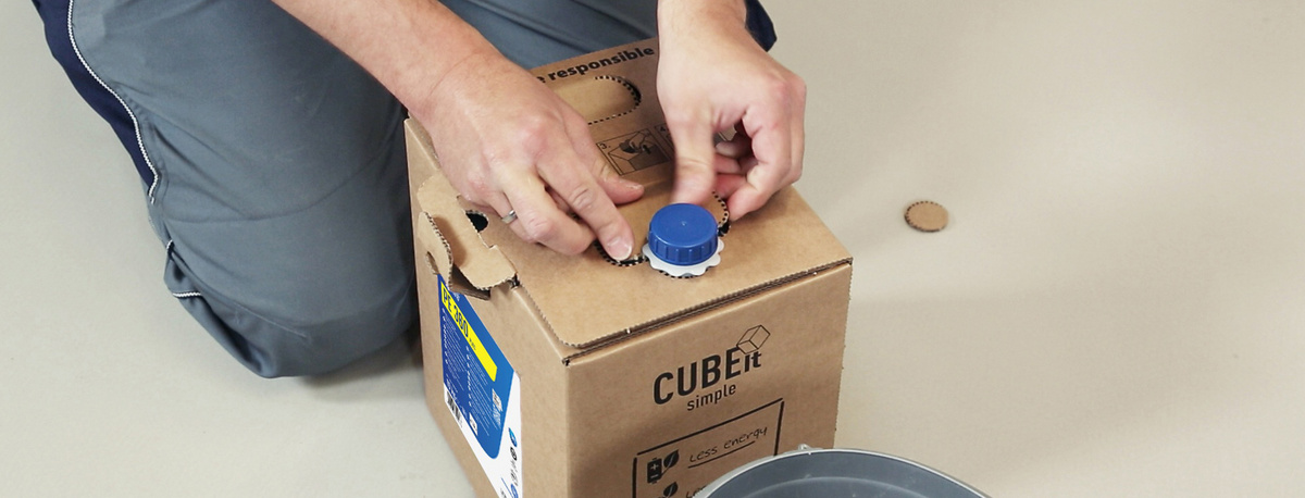 Eine Person handhabt eine CUBEit Verpackungseineinheit. Aus dem kubischen Pappkarton schaut oben die Ausschüttöffnung heraus.