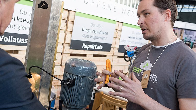Ein Mitarbeiter einer Repair-Initiative befindet sich im Gespäch mit einem Interessenten. Im Hintergrund sind Elemente einer Werkstatt zu erkennen.