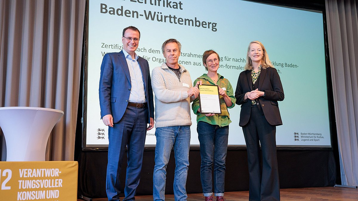 Umweltministerin Thekla Walker MdL und Kultusstaatssekretär Volker Schebesta MdL stehen zusammen mit einer Frau und einem Mann auf einer Bühne. Sie lächeln alle in die Kamera, die Frau und der Mann halten ein eingerahmtes BNE-Zertifikat.