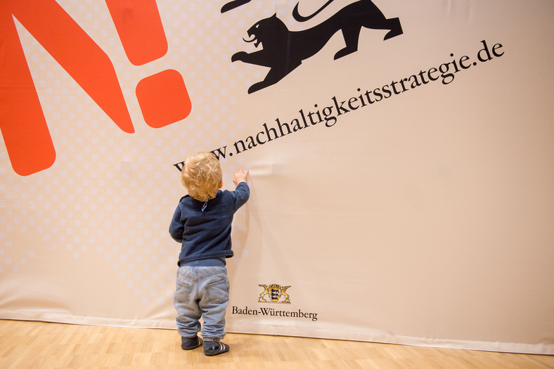Ein einjähriges Kind sieht man von hinten. Es berührt ein Plakat oder Banner, auf dem www.nachhaltigkeitsstrategie.de steht und das orangefarbene N.