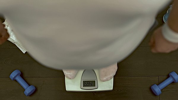 Dargestellt ist eine übergewichtige Person, die auf einer Waage steht. Daneben liegen Hanteln auf dem Boden.