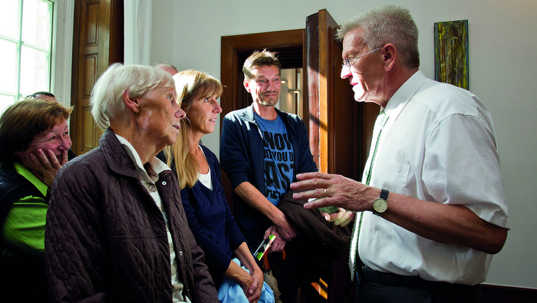 Offene Tür beim Ministerpräsidenten Baden-Württemberg Ministerpräsident Winfried Kretschmann erklärt einer Gruppe von sieben Leuten etwas in einem hellen Raum.