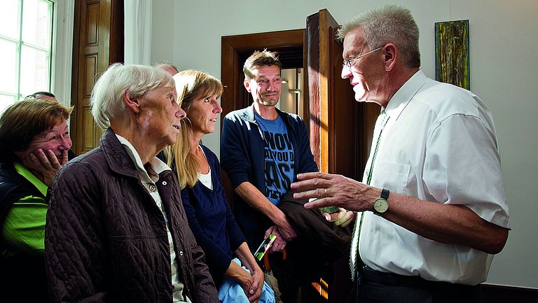 Ministerpräsident Winfried Kretschmann erklärt einer Gruppe von sieben Leuten etwas in einem hellen Raum. 