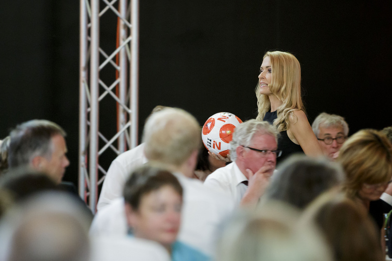 Die Moderatorin inmitten des sitzenden Publikums. Sie hält einen Lederfußball mit Nachhaltigkeitslogo in der Hand.