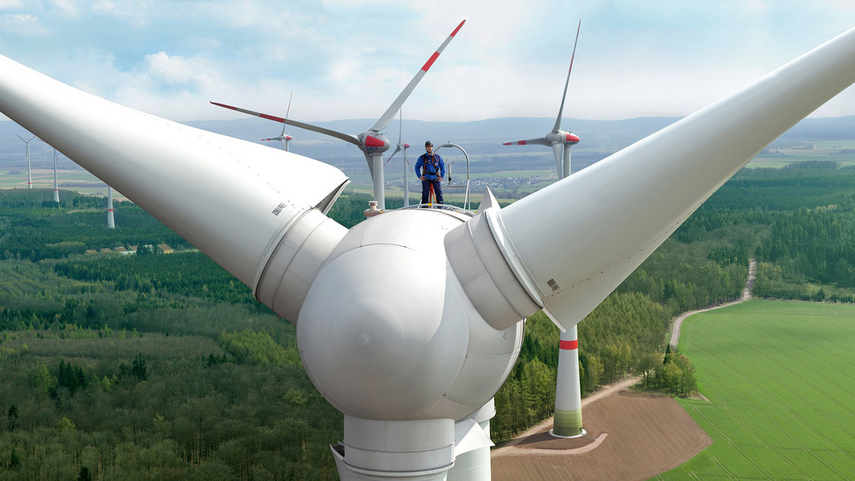 Groß dargestellt wird von einer Windkraftanlage die Gondel mit Rotorblättern. Ein Mitarbeiter steht oben auf der Gondel, im Hintergrund stehen weitere Windkraftanlagen.