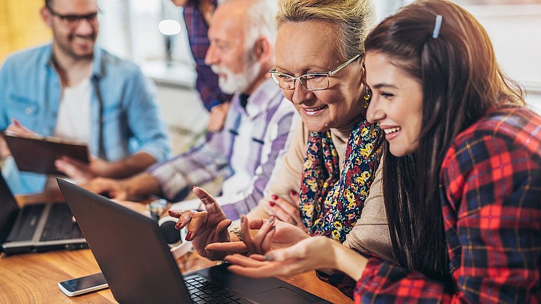 Eine jüngere und ältere Frau, die hinter einem Laptop sitzen und diskutieren. Im Hintergrund sind drei weitere Erwachsene verschiedenen Alters hinter einem Laptop zu sehen.