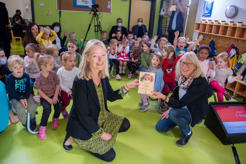 Thekla Walker MdL und eine Erzieherin zeigen den Preis in die Kamera. Im Hintergrund freuen sich die Kinder über den Gewinn