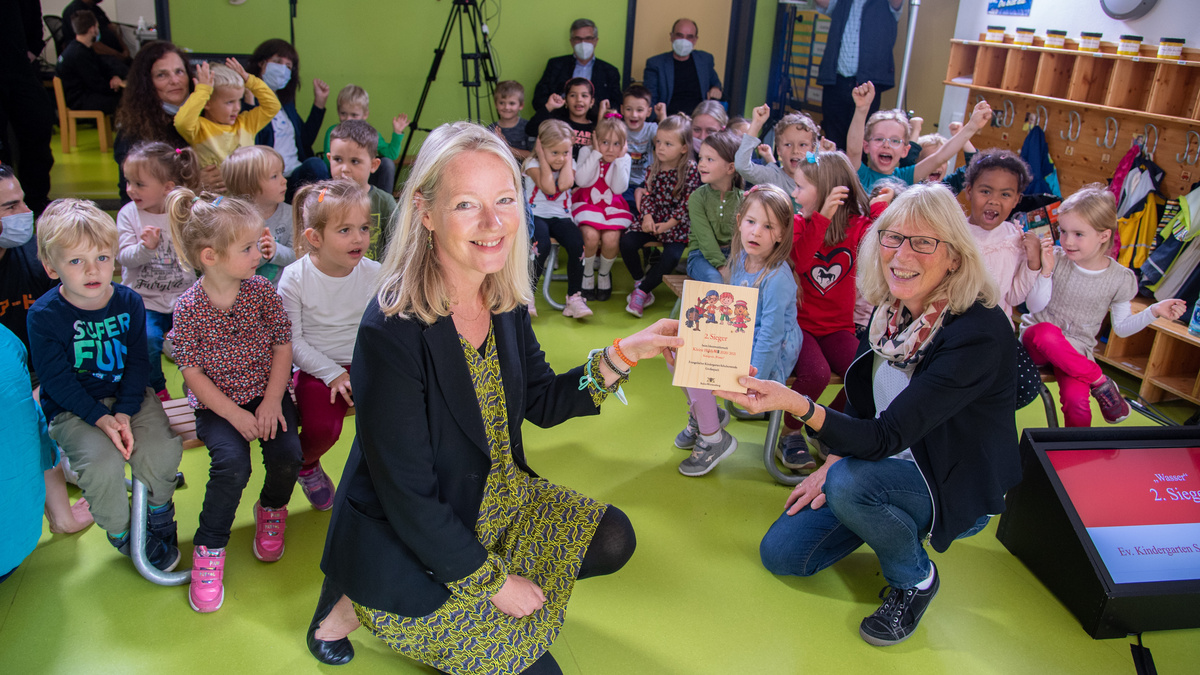 Thekla Walker MdL und eine Erzieherin zeigen den Preis in die Kamera. Im Hintergrund freuen sich die Kinder über den Gewinn