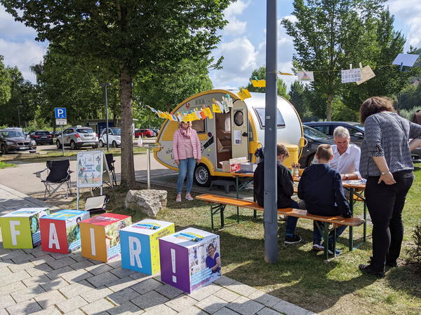 Ausstellung auf einem Parkplatz mit einem kleinen Wohnwagen und Würfeln mit der Aufschrift "fair".