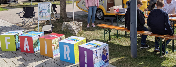 Ausstellung auf einem Parkplatz mit einem kleinen Wohnwagen und Würfeln mit der Aufschrift "fair".