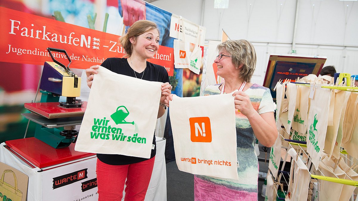 Zwei Frauen am Stand der Jugeninitiative Nachhaltigkeit Baden-Württemberg beim Kirchentag 2015 halten frisch bedruckte Baumwolltaschen von der Jugeninitiative Nachhaltigkeit Baden-Württemberg halten frisch bedruckte Baumwolltaschen hoch. 