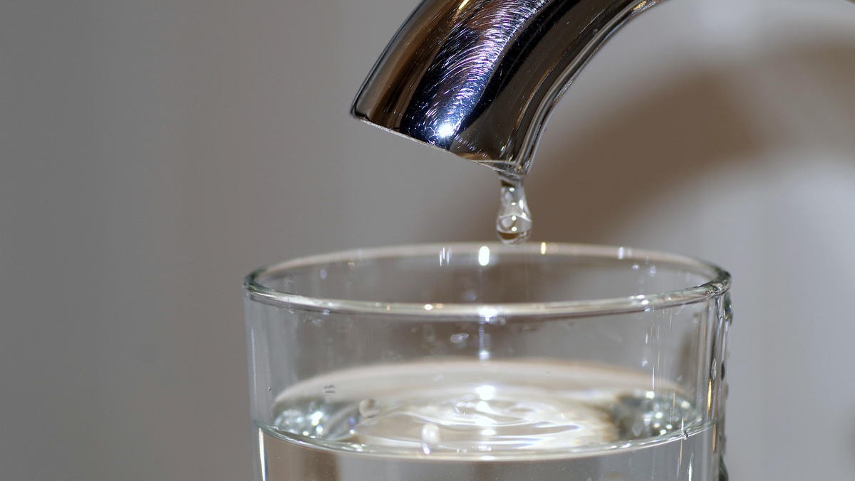 Aus einem Wasserhahn tropft ein Tropen Wasser in ein fast volles Wasserglas.