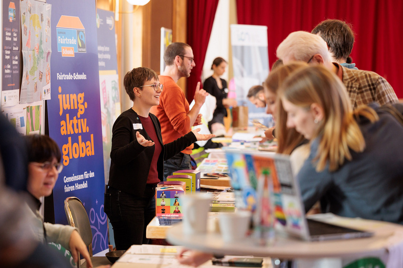 Das Farbfoto zeigt Menschen, die an Infoständen in einem Saal stehen. Eine Frau gestikuliert im Gespräch. Ein Plakat hinter ihr trägt den Text: „Fairtrade-Schools: jung. aktiv. global.“