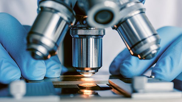 Dargestellt ist eine Person, die einen Objektträger unter das Objektiv eines Mikroskopes legt.