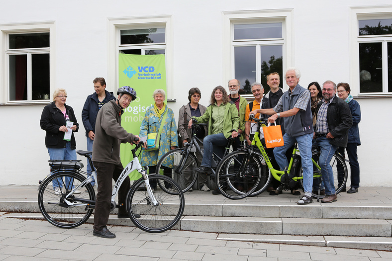Eine Gruppe von Menschen, drei mit Fahrrad vor einer Hauswand. Ein grünes Banner mit VCD-Logo im Hintergrund.