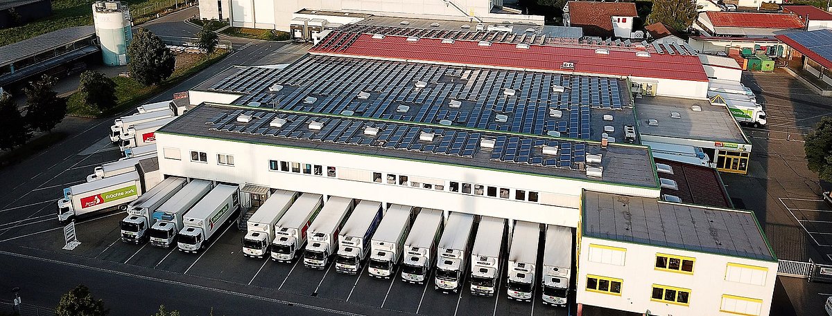 Eine Luftaufnahme des Firmengeländes der Früchte Jork GmbH. Auf den Dächern ist die Photovoltaik-Anlage erkennbar und im Hintergrund die Allgäuer Landschaft.