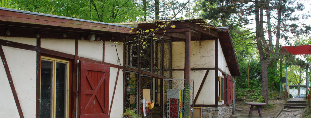 Ein Fachwerkhaus mit roten Fensterläden, ein Weg führt durch ein Tor. Im Hintergrund Bäume, vor dem Haus stehen alte Sitzmöbel.