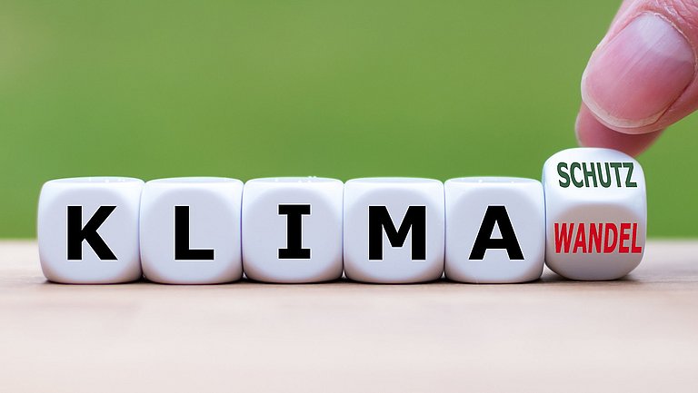 Fünf Spielwürfel mit Buchstaben bilden das Wort Klima. Auf einem letzen Würfel in der Reihe steht auf einer Würfelfläche das Wort Wandel und auf der anschließenden das Wort Schutz. Dieser Würfel wird von zwei Fingern auf Kippstellung zwischen Wandel und Schutz gehalten.