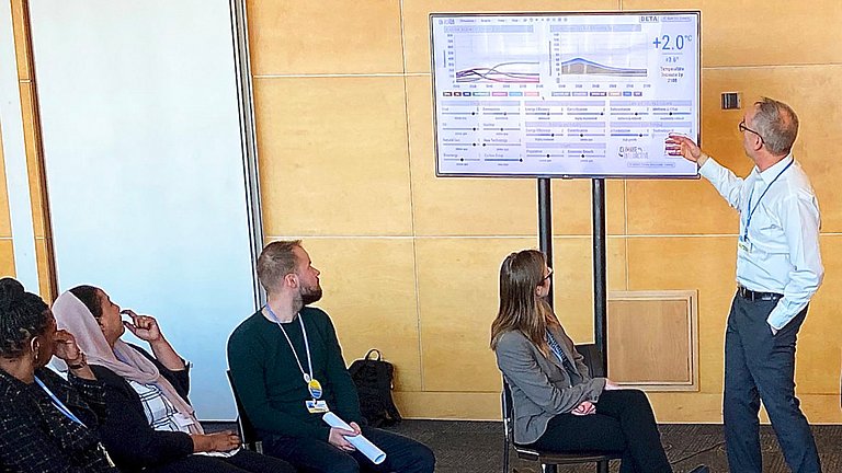 Mehrere Erwachsene, die im Stuhlkreis sitzen und dem Vortragenden Zuhören. Der Vortragende steht vor einem Bildschirm, auf dem seine Präsentation zu sehen ist.