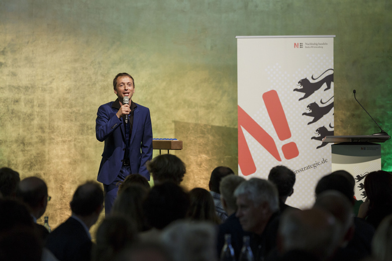 Ein Mann im blauen Anzug auf der Bühne spricht oder singt ins Mikrofon.