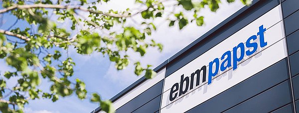 Ansicht auf das Werksgebäude von ebm-papst mit Fokus auf das Logo, links ist ein grüner Baum zu sehen.